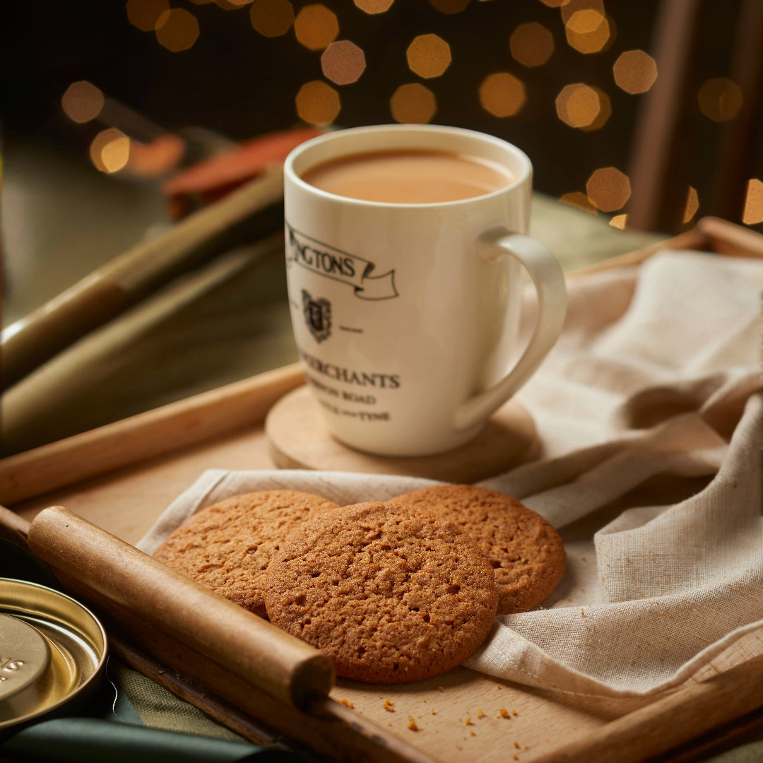 A white ceramic mug filled with a warm beverage sits on a wooden tray beside two round cookies on a beige cloth, with a rolling pin and soft warm lights blurred in the background.