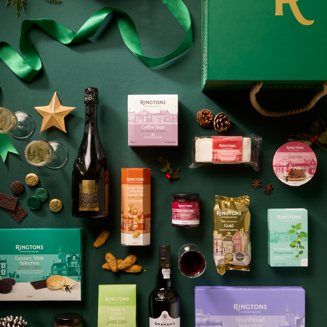A festive flat lay of Ringtons luxury gift basket contents on a dark green background, featuring a bottle of sparkling wine, a bottle of Graach wine, assorted chocolates, biscuits, a cheese block, and gift boxes, alongside a green ribbon and a green gift bag.