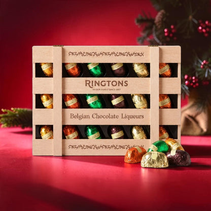 A wooden gift box of Ringtons Belgian Chocolate Liqueurs chocolates, with several wrapped chocolates placed in front on a red surface against a dark red background with a blurred Christmas tree.