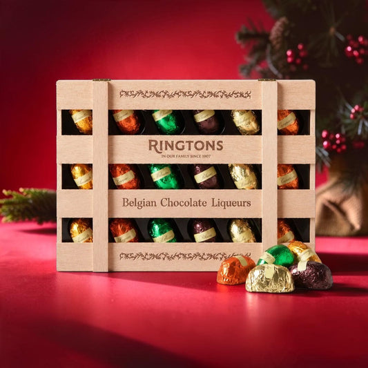 A wooden gift box of Ringtons Belgian Chocolate Liqueurs chocolates, with several wrapped chocolates placed in front on a red surface against a dark red background with a blurred Christmas tree.