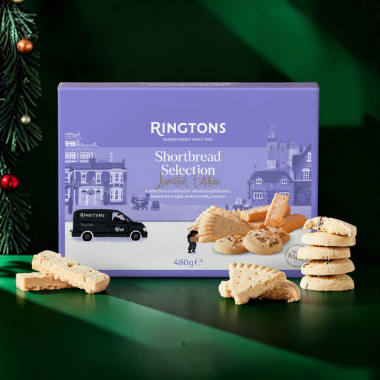 Ringtons Shortbread Selection Limited Edition box of cookies displayed on a dark green surface with Christmas tree branches and ornaments at the left edge. Broken and stacked shortbread biscuits are arranged around the box.