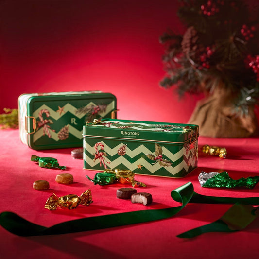 Two green Juncions brand chocolate tins with white chevron patterns and holiday illustrations, displayed on a red surface with scattered wrapped chocolates and a dark festive tree in the background.