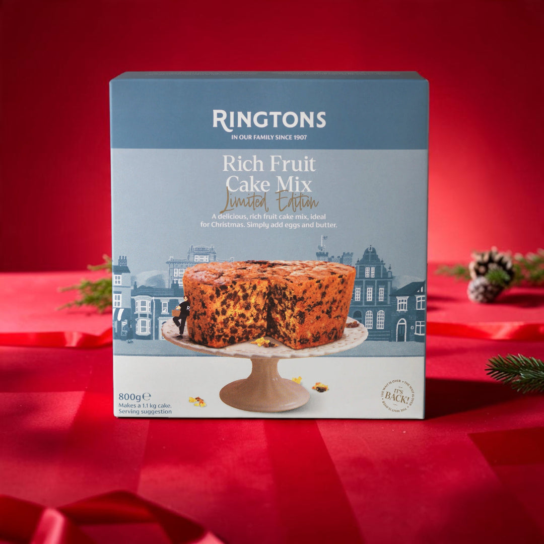 A boxed cake mix product standing upright on a red satin surface. The package is blue and white with the brand name Ringtons and the product name Rich Fruit Cake Mix, plus a photo of a sliced moist cake on a stand.
