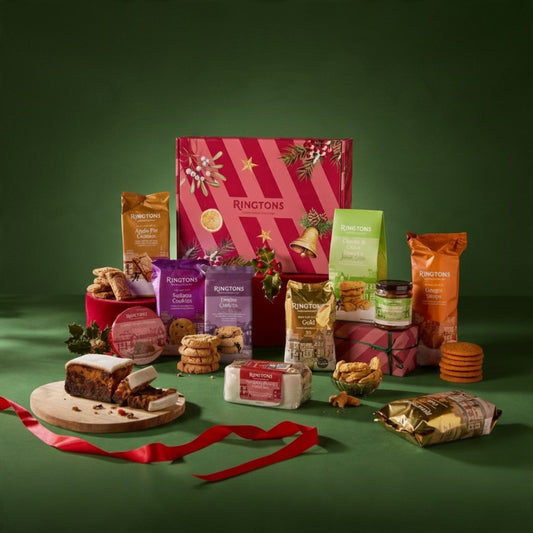 A Ringtons Christmas gift hamper arranged on a dark green background, featuring a large pink striped gift box with festive illustrations, assorted packaged snacks and biscuits, a sliced Christmas pudding on a wooden board, cookies, a jar, and red ribbon accents.