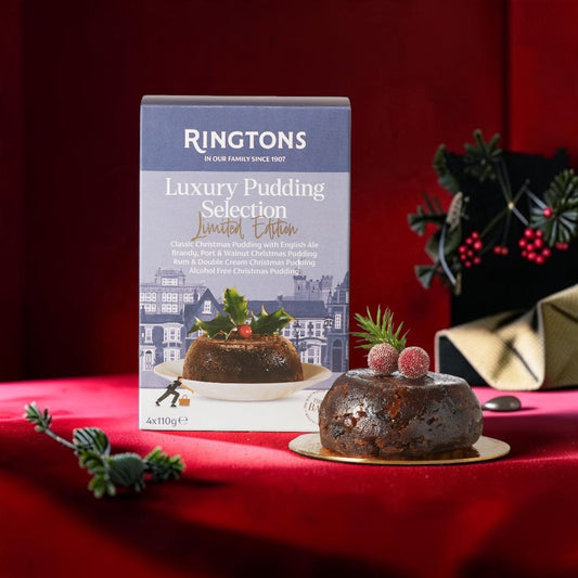 Ringtons Luxury Pudding Selection Limited Edition box displayed next to a decorated Christmas pudding on a red fabric surface with holiday greenery and a gold napkin in the background.