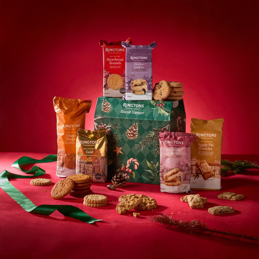 A Ringtons biscuit gift hamper set against a deep red background, featuring a green pine-patterned box surrounded by assorted biscuit bags in orange, gold, pink, and white, with cookies scattered around.