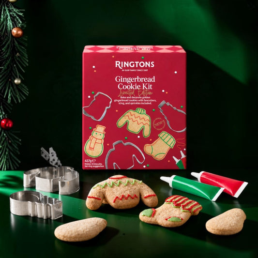 Ringtons Gingerbread Cookie Kit box displayed with metal cookie cutters, raw gingerbread pieces, and two icing tubes on a dark green surface with a Christmas tree branch at the edge.
