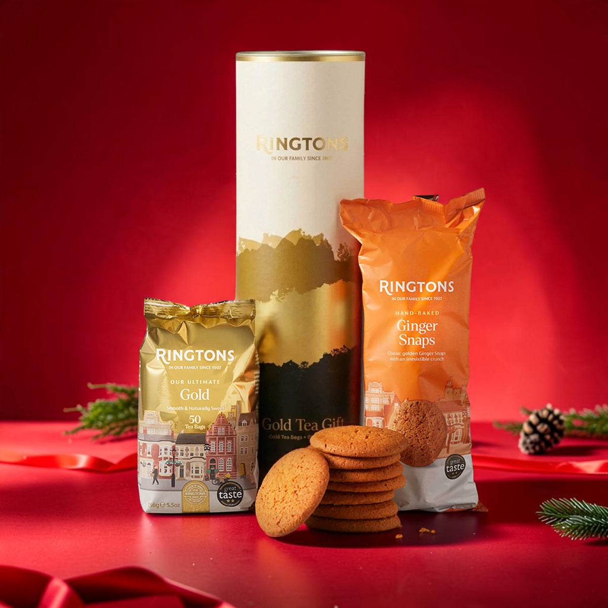 Ringtons Christmas gift set featuring a tall white and gold cylindrical tin, a gold foil bag of Gold Tea Gift cookies, and an orange bag of Ginger Snaps cookies, arranged on a red surface with red ribbon and holiday greenery against a red background.