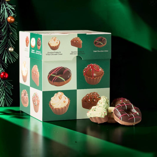 A boxed assortment of frosted donuts and cupcakes sits on a glossy dark green surface with a matching green backdrop. The box is white and green with printed images of assorted pastries and a small pull tab.