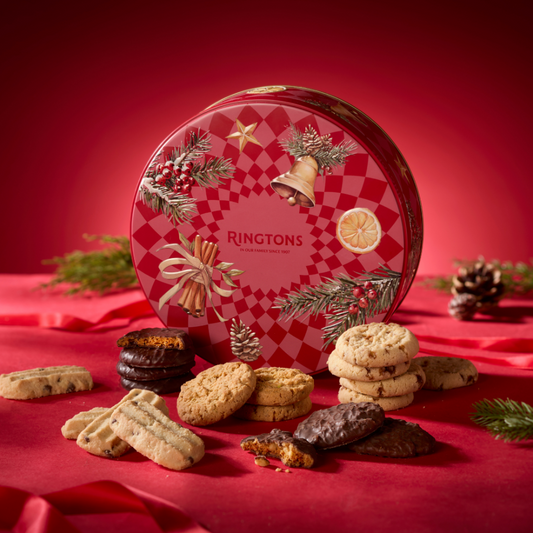 A festive round tin of Ringtons Christmas Cookie Bar displayed with assorted cookies on a red fabric surface against a red background. The tin features a red-and-white checkered pattern with illustrations of a bell, star, cinnamon stick, and lemon slice.