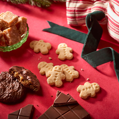 Gingerbread man cookies arranged on a red surface with chocolate pieces, a green glass bowl of brown sugar cubes, and holiday ribbon props.