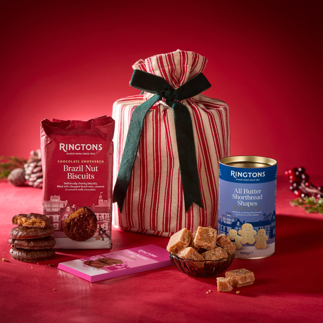 A holiday gift hamper of Ringtons biscuits arranged on a red surface against a red background. The set includes a large red-and-white striped gift bag tied with a dark green ribbon, a bag of Ringtons Brazil Nut Biscuits, a can of Ringtons All Butter Shortbread Shapes, a pink chocolate bar, a bowl of sugar cookies, and a stack of chocolate nut cookies.